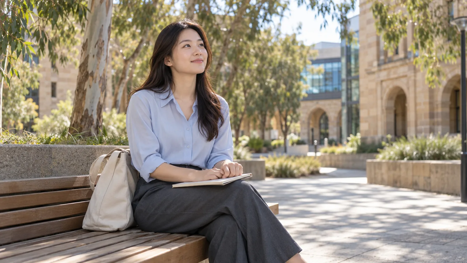 A Malaysian student sits with a notebook on a bench in a sunlit Australian university courtyard.
